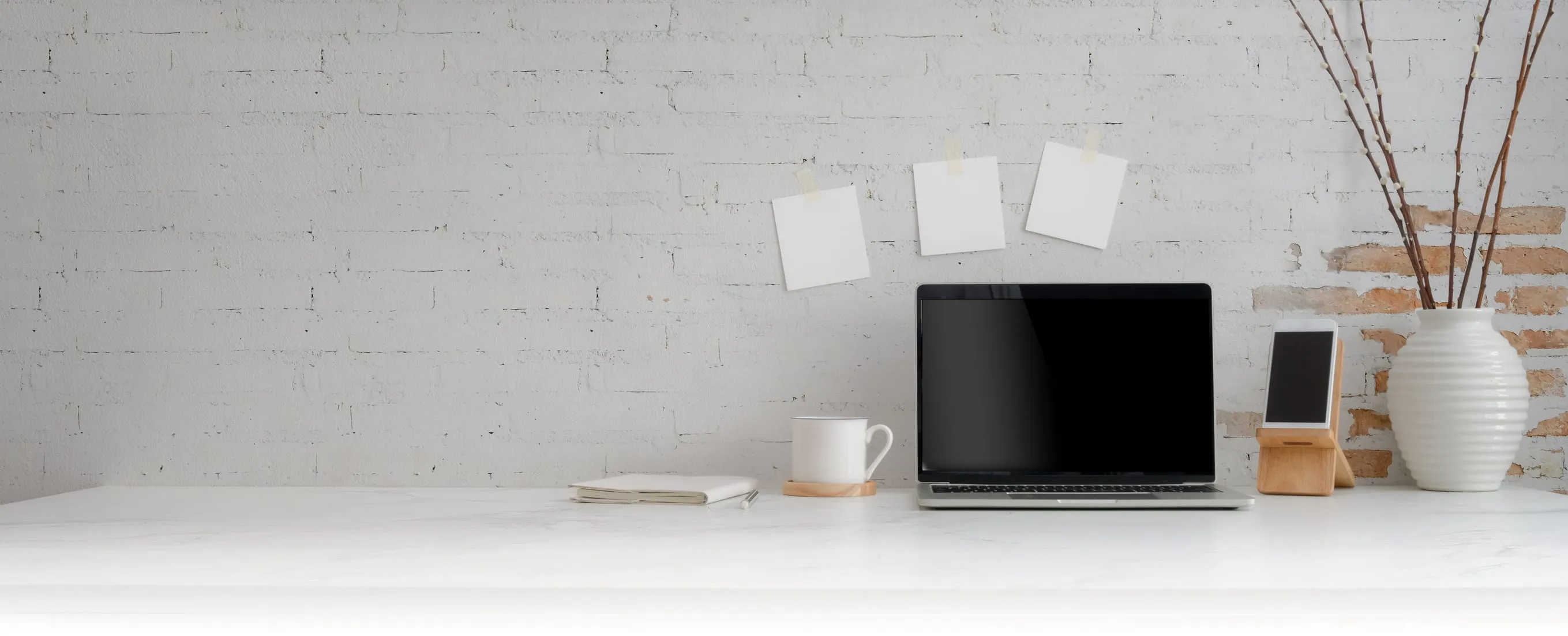 A calm home-office desk with a laptop, notebook and coffee against a whitewashed brick wall