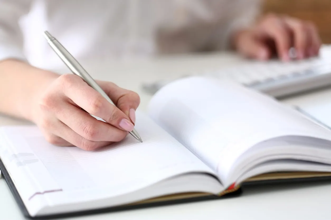 A hand writing in a paper diary beside a keyboard, planning the week ahead
